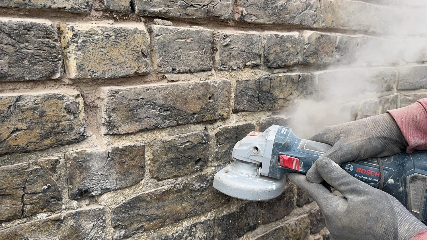 Angle grinder raking out an eroded mortar joint in London stock brick, dust rising from the cut