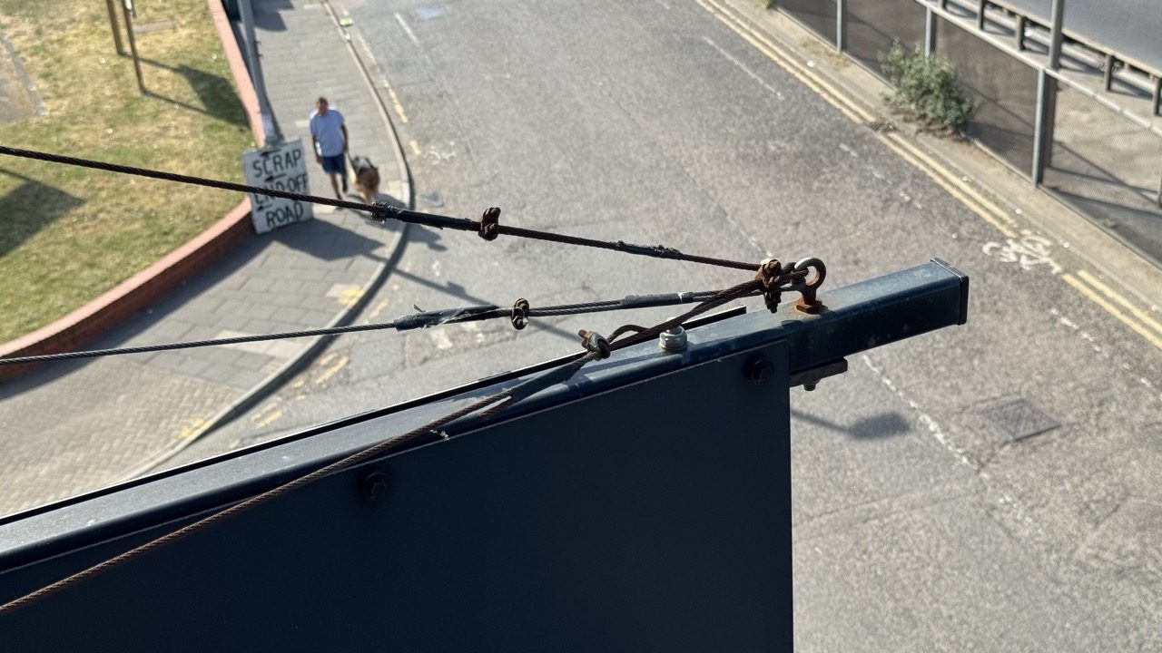 Corroded wire rope retention fitting at the top of a sign gantry, seen from above with the road below